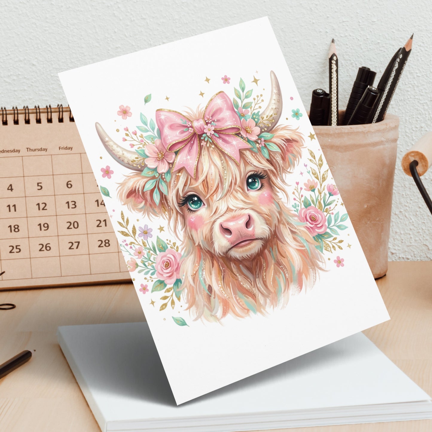 Watercolor highland cow with floral accents and pink bow on card.
