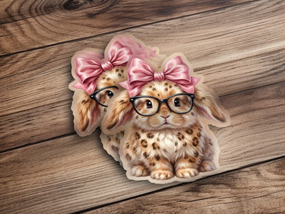 Two spotted bunnies with pink bows and glasses on a wooden background.