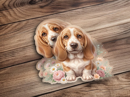 Two basset hound puppies sit surrounded by flowers on a wooden background.