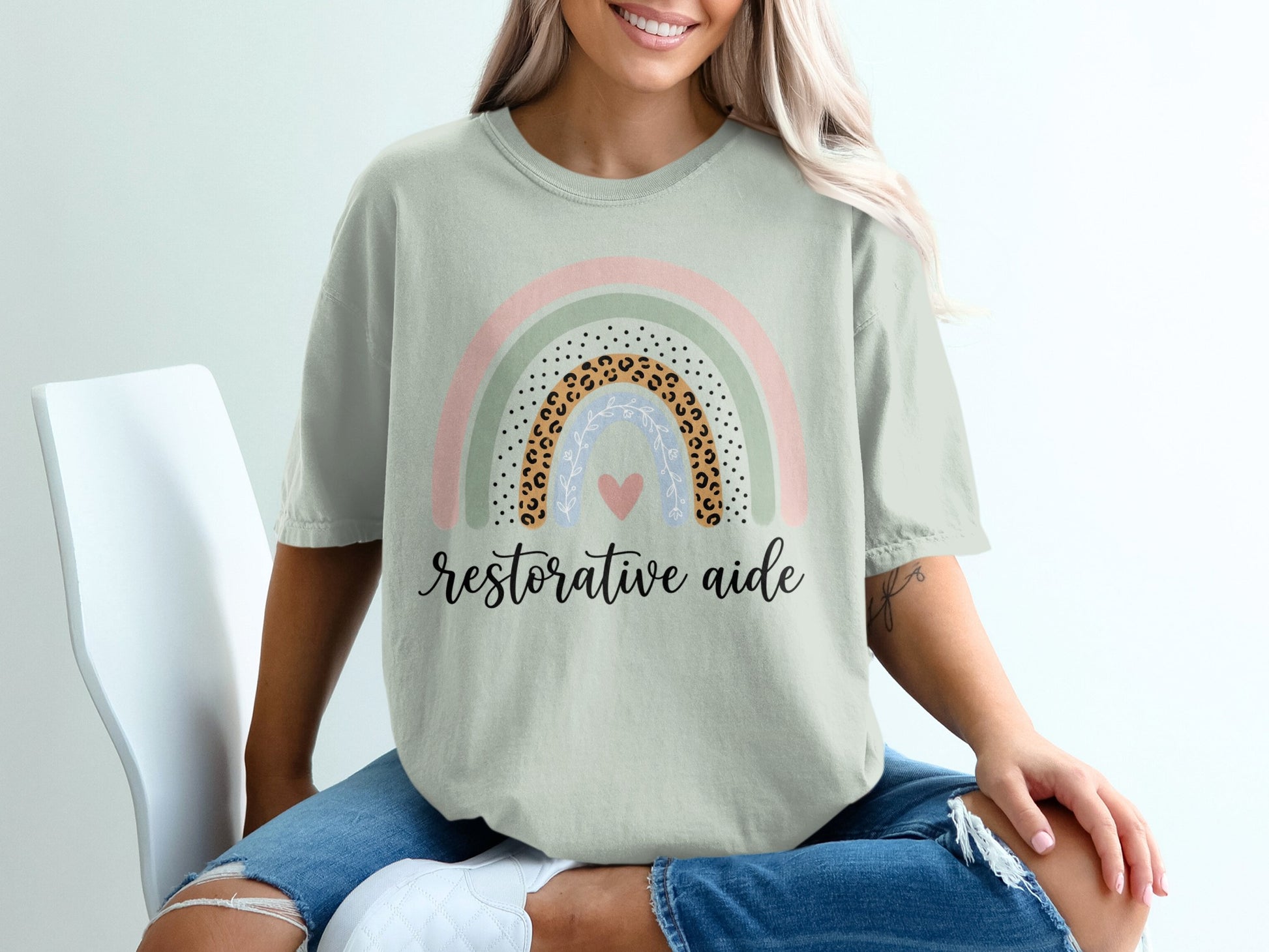 Light sage green t-shirt with a pastel rainbow graphic and text "restorative aide".