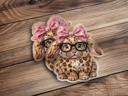 Two plush bunny patches with pink bows and glasses on a wooden background.