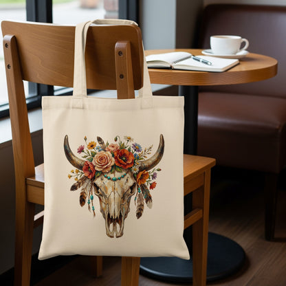 Cream tote bag with a colorful floral and feather-adorned bull skull graphic.