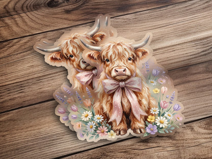 Two Highland cows with pink bows amidst wildflowers on a wooden background.