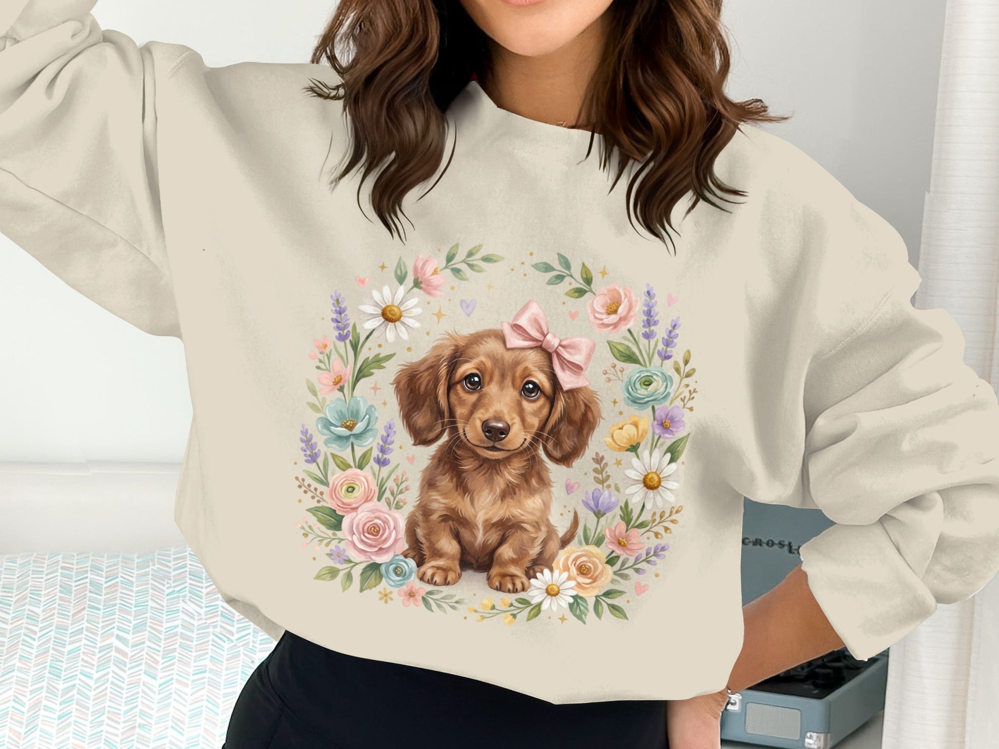 Dachshund puppy wearing pink bow surrounded by floral wreath on cream sweatshirt.