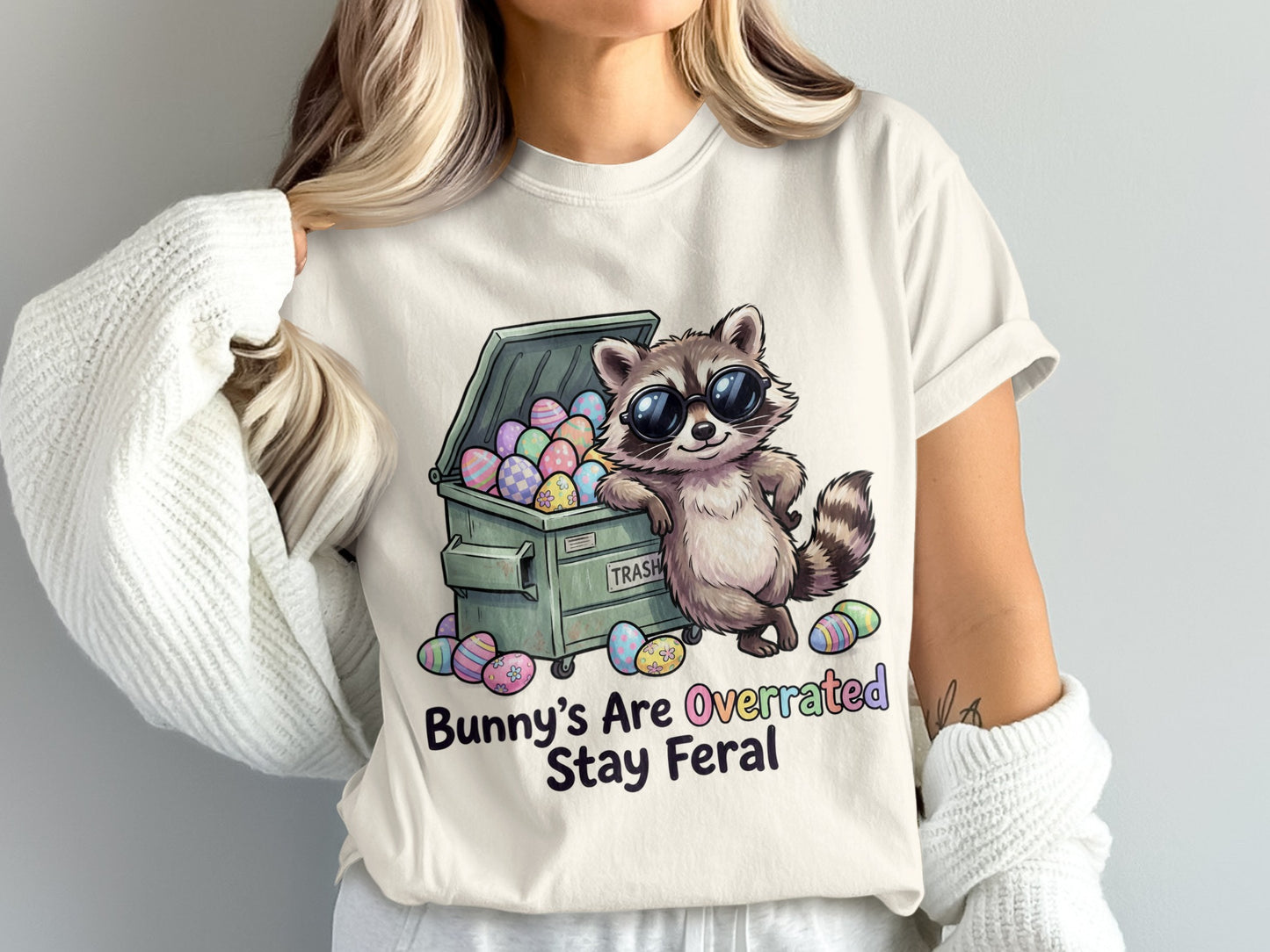 Raccoon in sunglasses leaning on a dumpster full of Easter eggs, text says "Bunnies are Overrated Stay Feral".