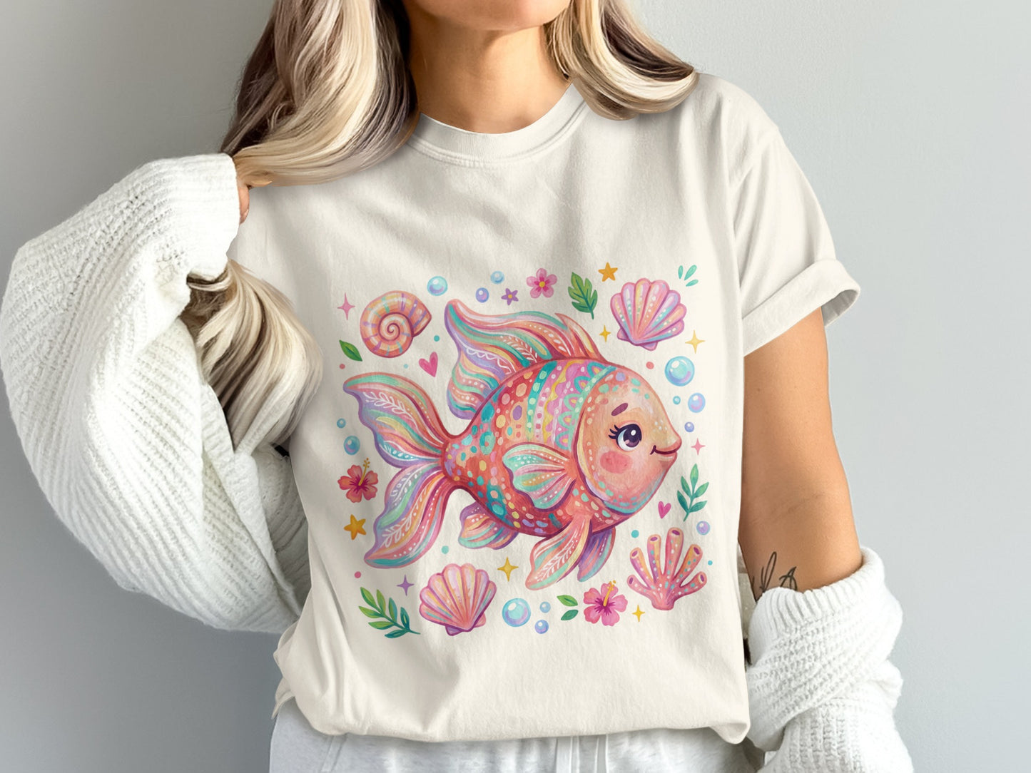 Woman wearing a cream t-shirt with a pastel goldfish and seashell graphic.