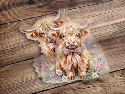 Two Highland cows with pink bows stand in a field of flowers.
