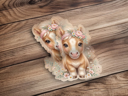 Two cute cartoon ponies wearing floral crowns on a wooden background.