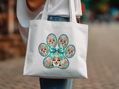 White tote bag with a paw print filled with corgi faces and a teal bow.