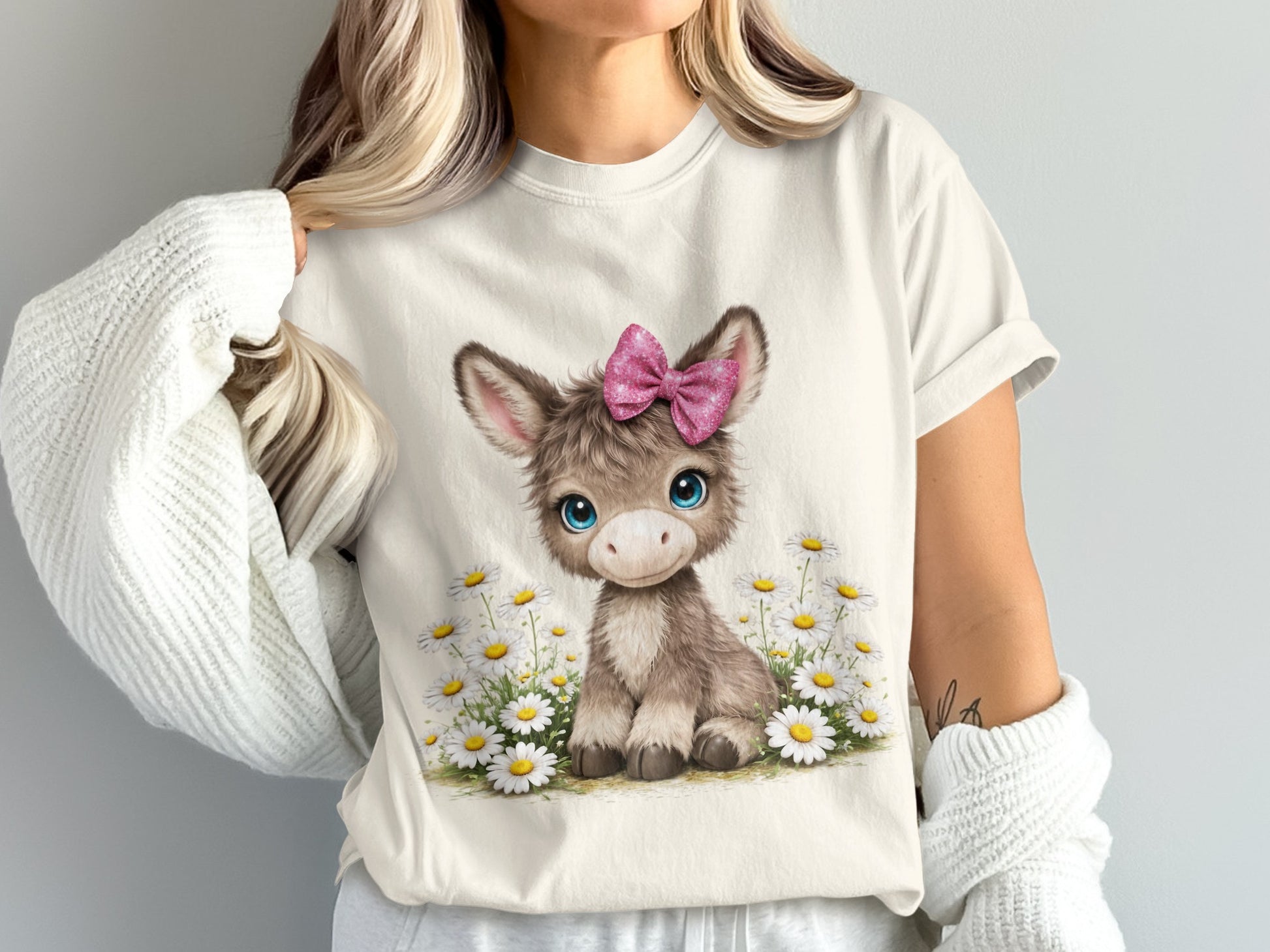 A fluffy baby donkey with a pink bow sits in a field of daisies.