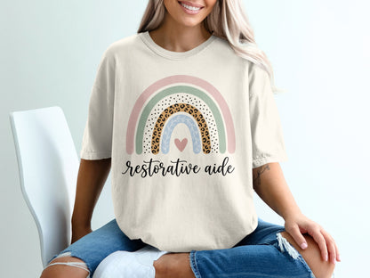Beige t-shirt with pastel rainbow and "restorative aide" text.
