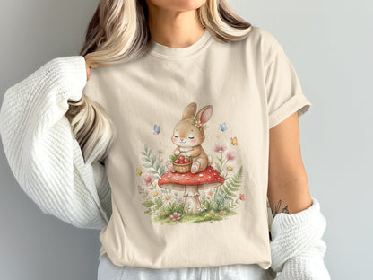 Cream t-shirt with a watercolor illustration of a bunny on a mushroom with flowers and butterflies.