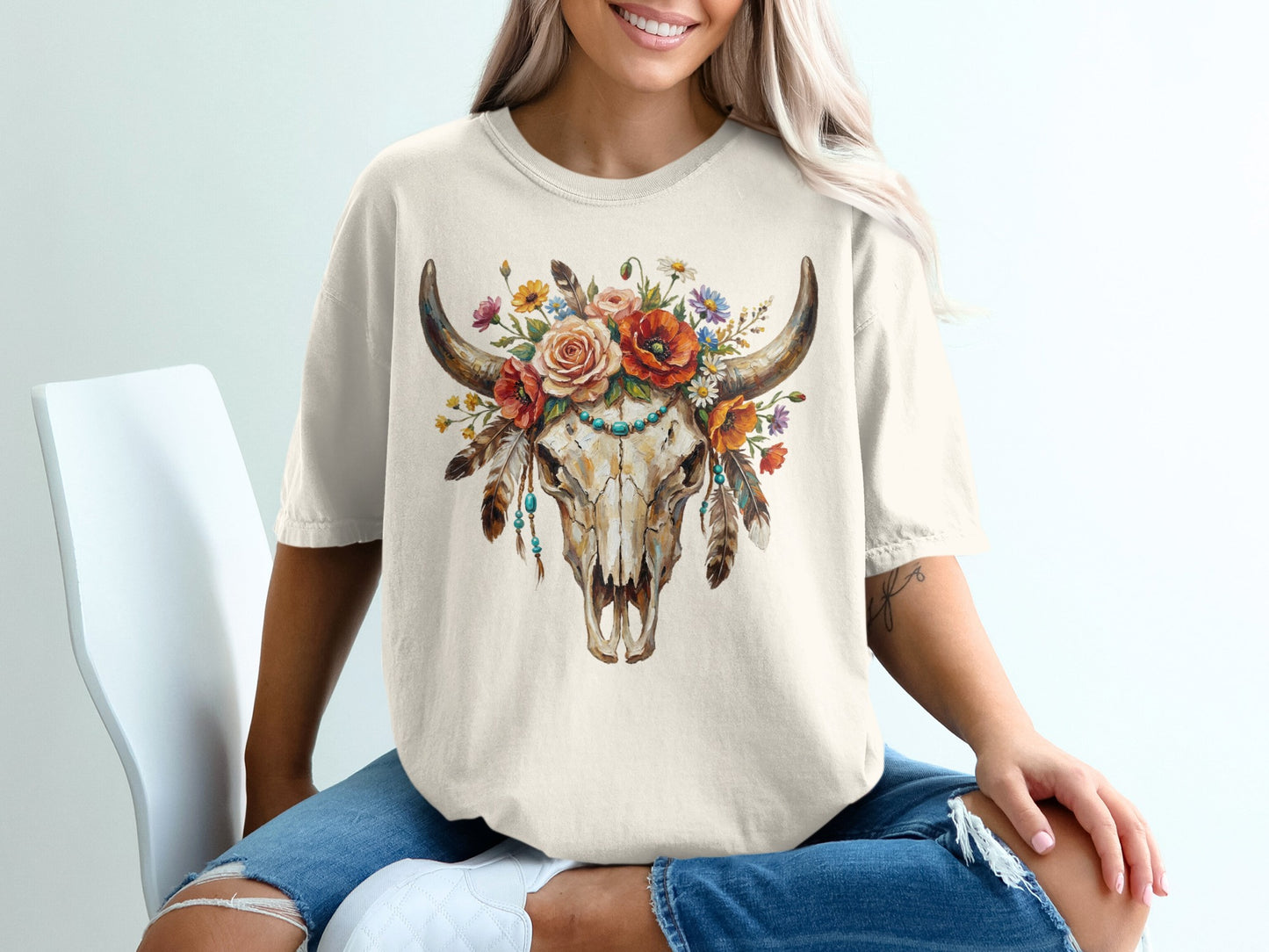 Cow skull with flowers and feathers graphic on cream t-shirt.
